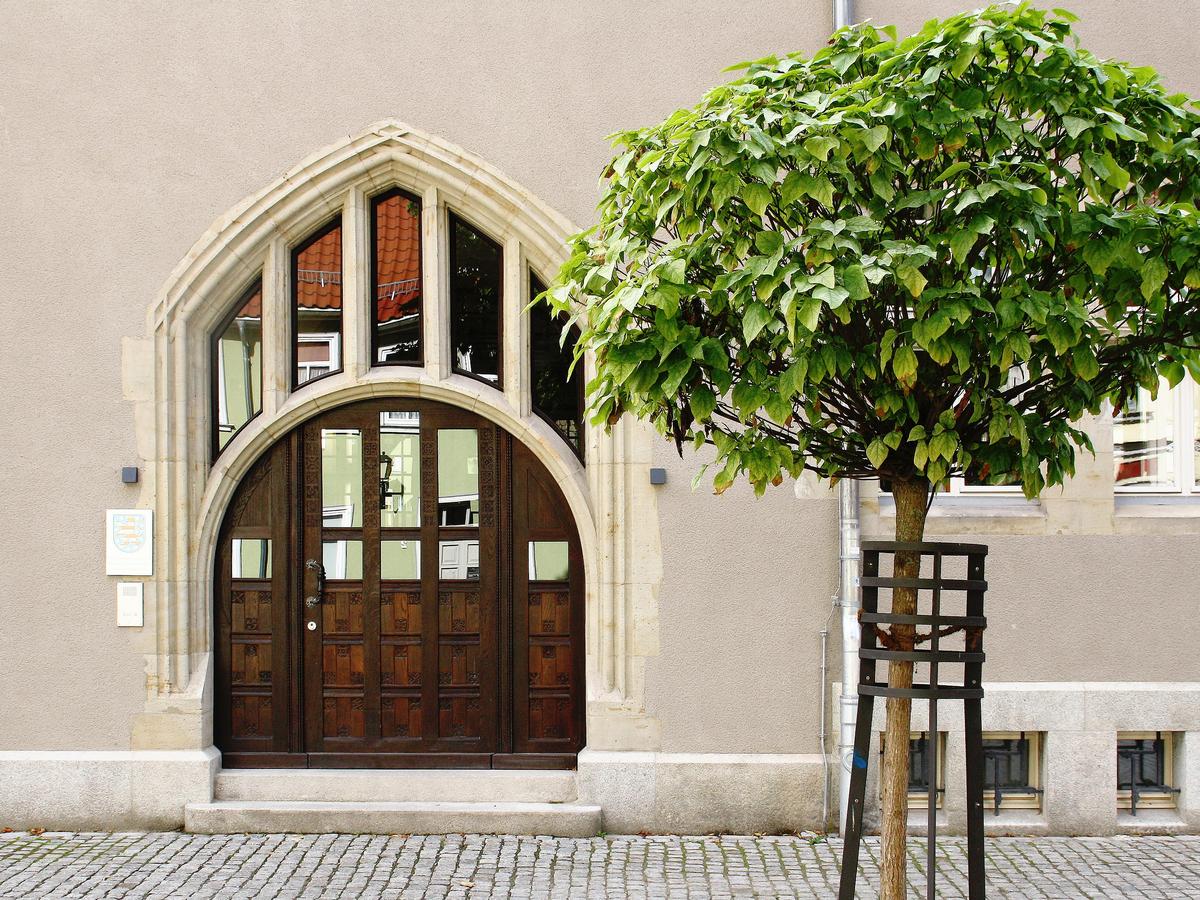 Detailfoto der Fassade mit einem jungen Baum mit kreisrunder Krone im Vordergrund. Die Tür mit Torbogen aus verziertem, durch die Zeit verdunkelten, Holz ist geschlossen und hat etliche Glasausschnitte. In dem Türbogen säulenartige Darstellung mit Glaselementen dazwischen.