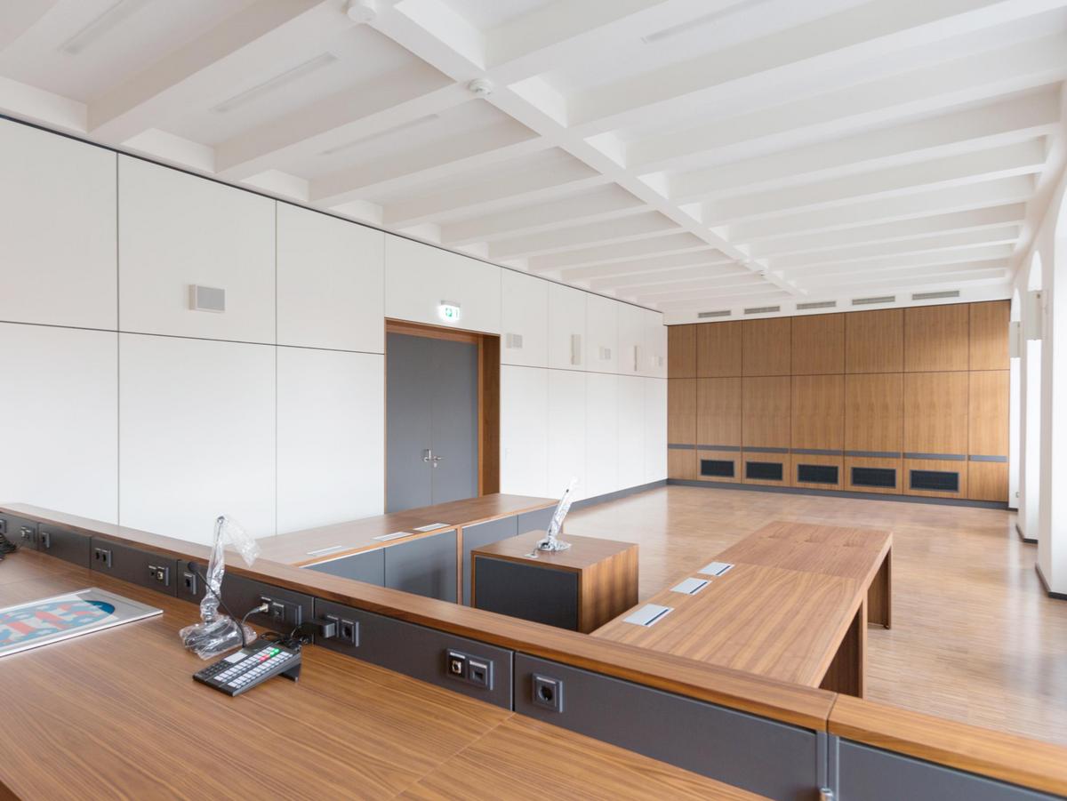 Großer Raum mit weißen Wänden und Holz-Stirnwand. Graue große zweiflügelige Eingangstür in der Mitte mit Holzzarge. Im Vordergrund der Richtertisch. Alle horizontalen Möbelbauteile in Holz, alle vertikalen Möbelbauteile in anthrazit. Hinter dem Richtertisch zwei Tische für die Gegnerparteien um 90° abgewinkelt und ein kleiner Tisch in der Mitte.
