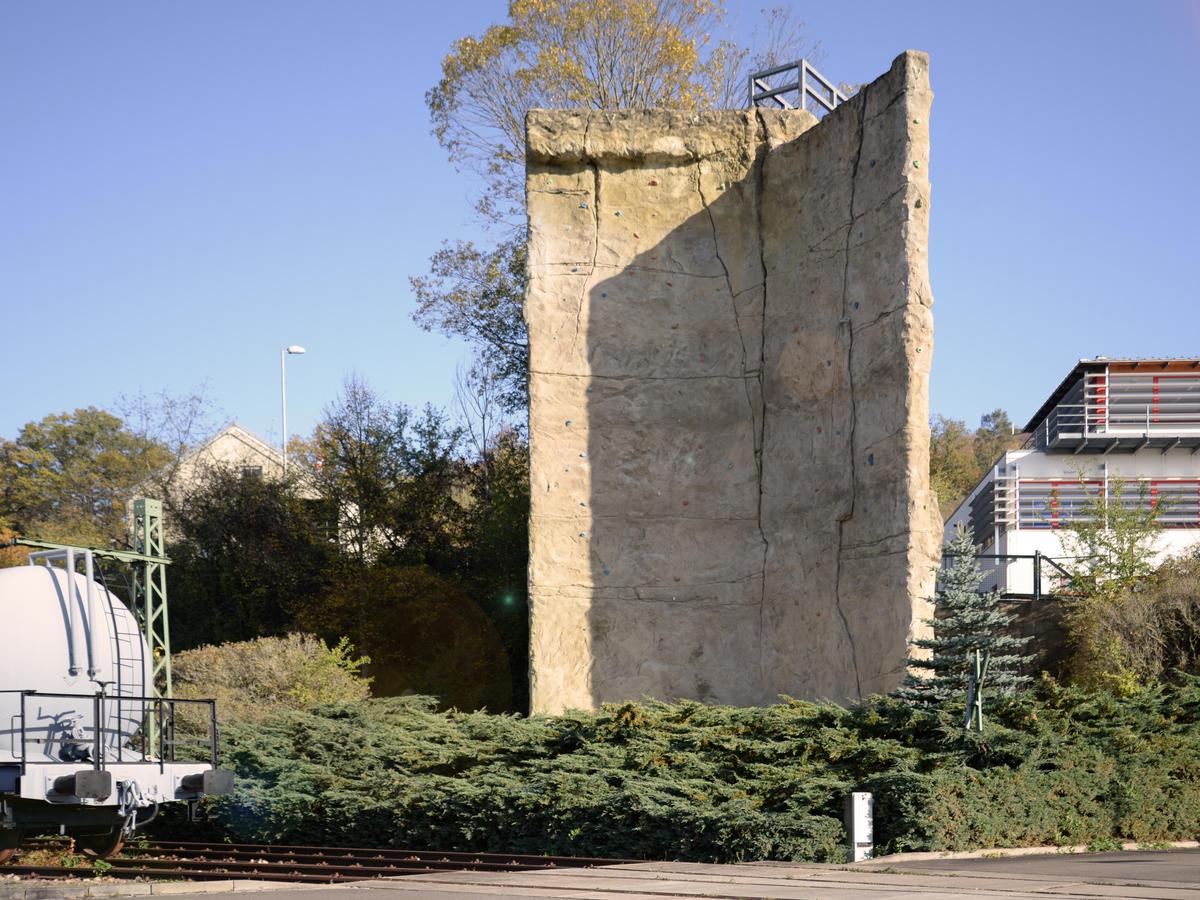 Übungs-Kletterturm halb im Schatten, mit Baum im Hintergrund als Maßstab. Im Vordergrund eingerahmt durch Grünpflanzen. Links angeschnitten sieht man den Übungs-Eisenbahnwaggon. Im Hintergrund die Brandsimulationsanlage.