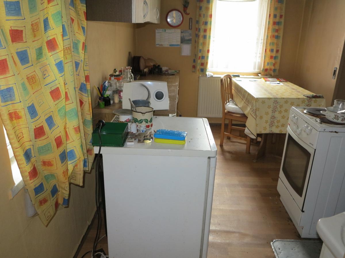 Blick in schmale Küche mit zwei Fenstern (an zwei Wänden über Eck); Linoliumfußboden ist gewellt; ein E-Herd und ein Kühlschrank, eine kurze Küchenzeile und eine Tisch stehen in der Küche