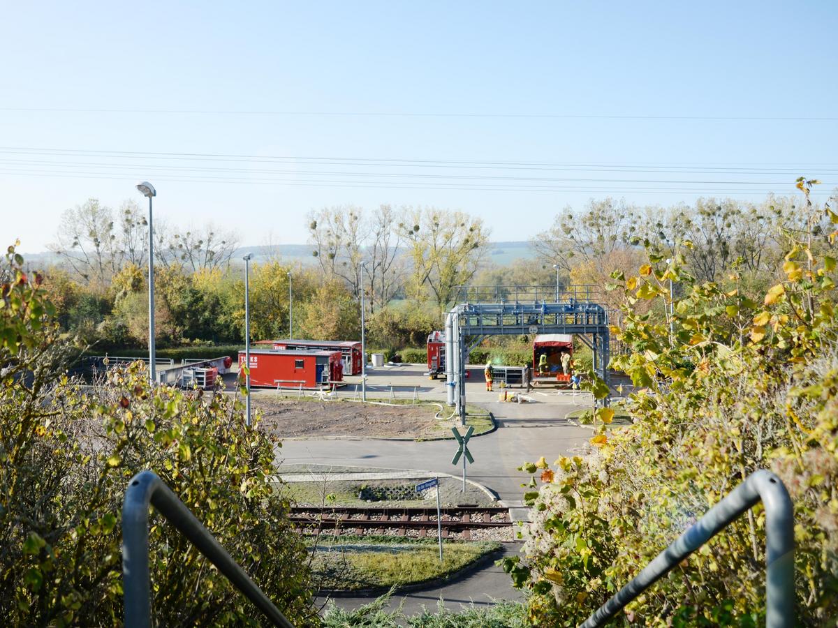 Blick auf den Übungsplatz mit Übungs-Leitungsbrücke, Feuerwehrfahrzeuge entladen Ihre Werkzeuge, an Bildrändern Sträucher, Container mit Ausrüstung der Feuerwehr stehen auf dem Übungsplatz.