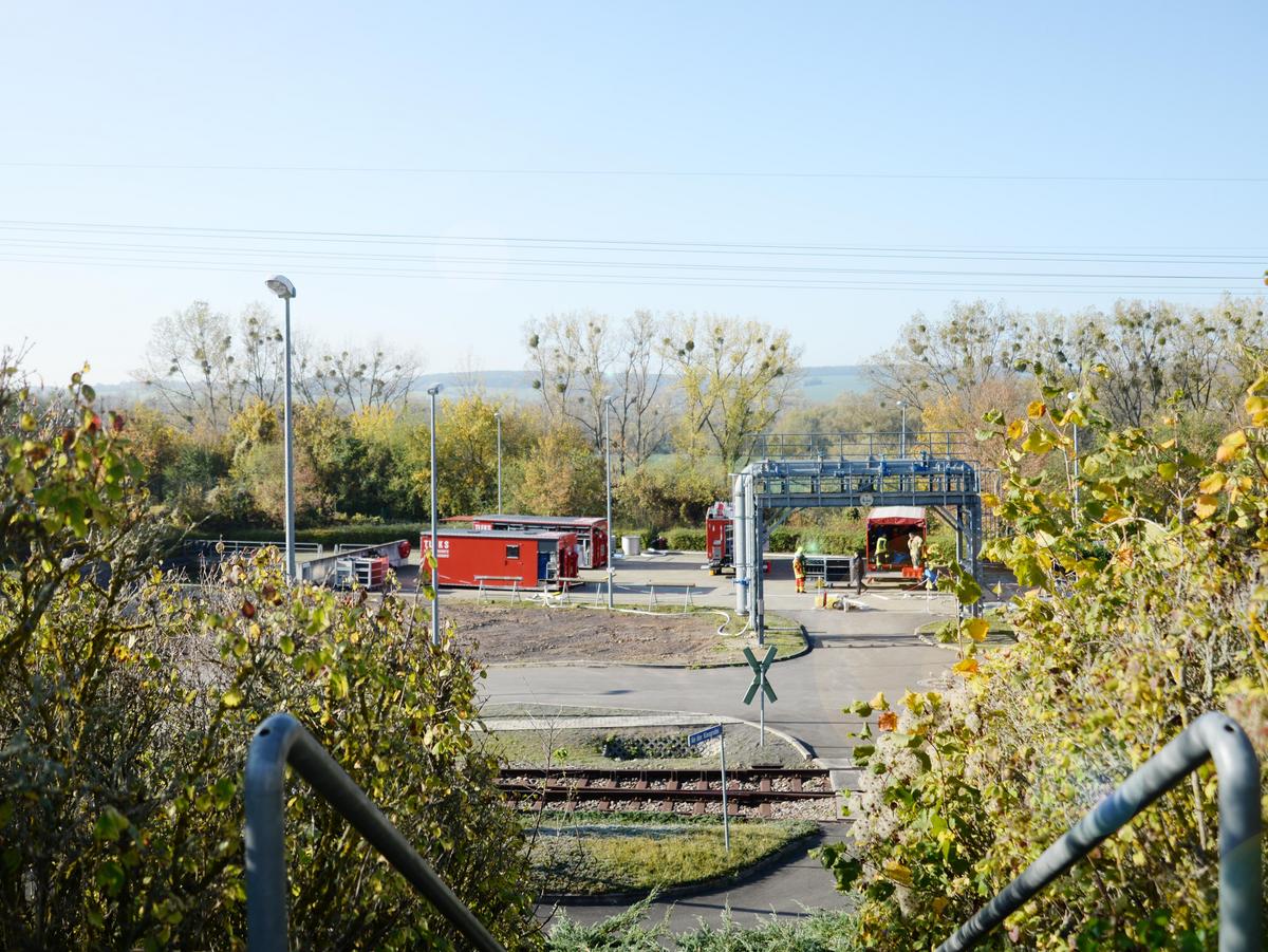 Blick auf den Übungsplatz mit Übungs-Leitungsbrücke, Feuerwehrfahrzeuge entladen Ihre Werkzeuge, an Bildrändern Sträucher, Container mit Ausrüstung der Feuerwehr stehen auf dem Übungsplatz.