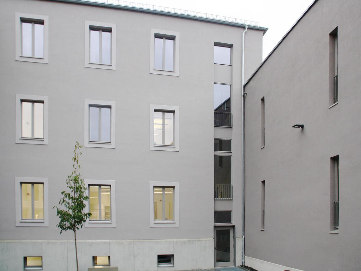 Foto der Lochfassade des Neubaus von der Hofseite. Eine graue Fassade mit einem jungen Baum davor. Die Fassade nimmt das Bild komplett ein, man sieht kaum Himmel und nur sehr wenig graues Pflaster im Vordergrund.
