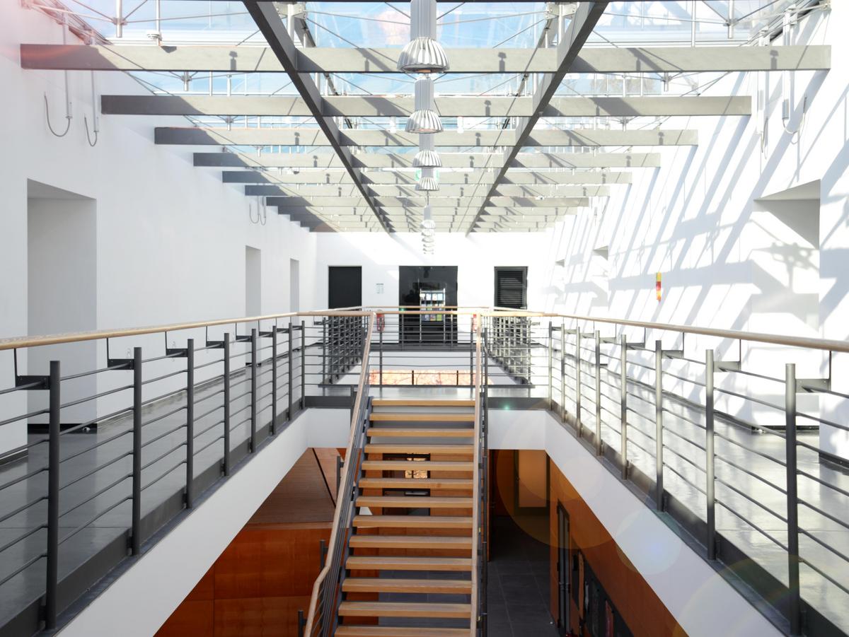 Atrium mit umlaufenden Galeriegang, im Luftraum eine Lusttreppe mit Holzbelag. Alles ist hell, lichtdurchflutet, transparent und freundlich. Das Dach ist komplett verglast.