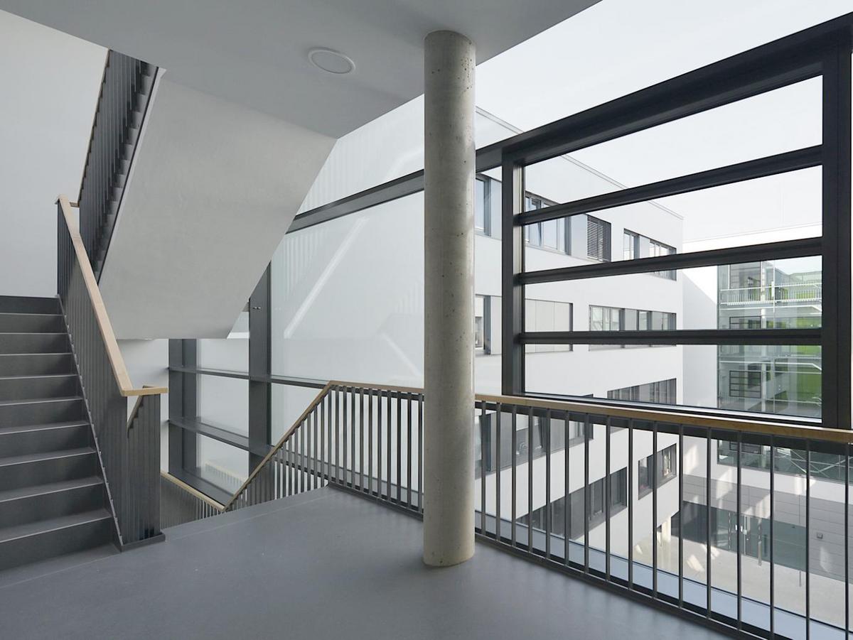 Treppenhaus links angeschnitten, Bild mit prominenter Betonstütze in Bildmitte. Es ist eine großzügige Verglasung zu sehen, durch die man in den Innenhof schaut. Weiße Fassaden mit Fensterbändern sind zu sehen, sowie der blaue Himmel.