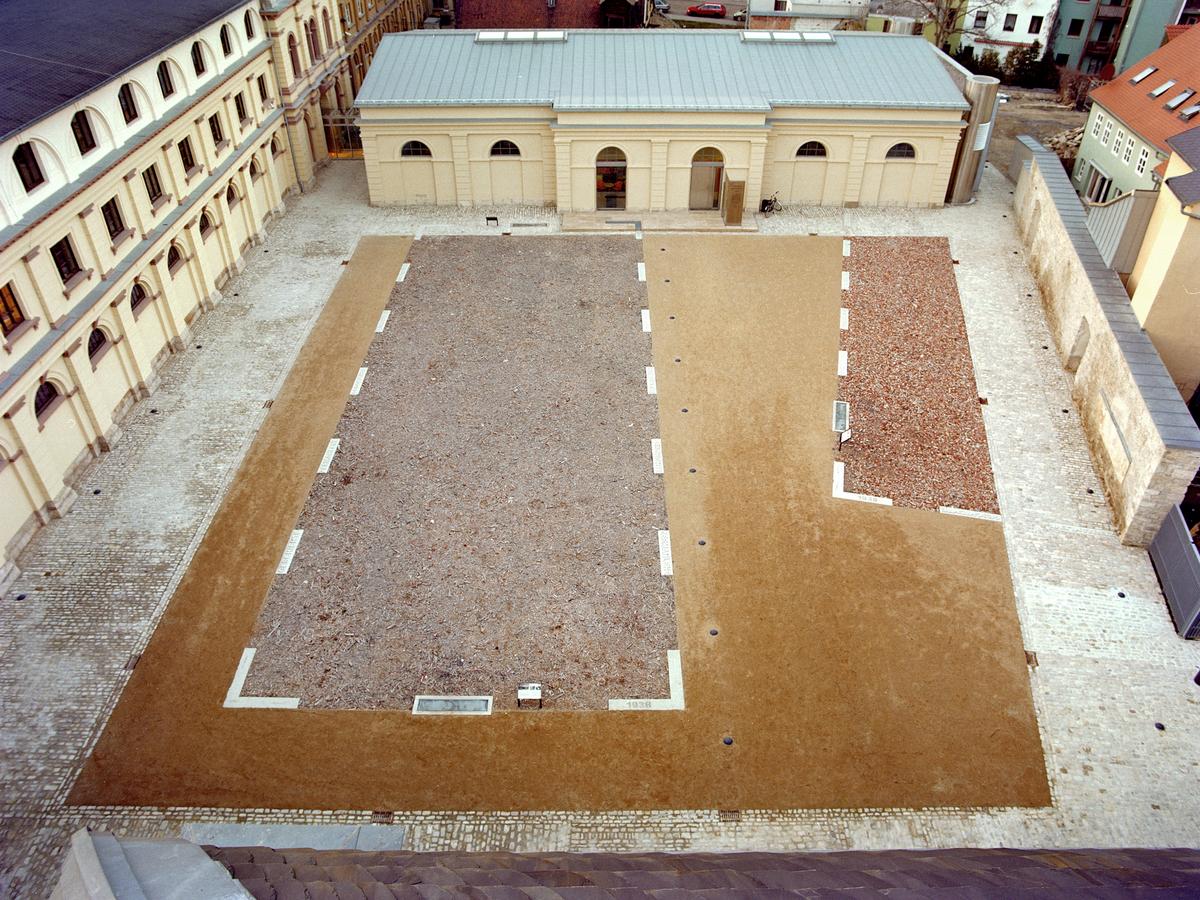 Blick vom Mittelrisalit auf den südlichen Marstallhof. Der Innenhof ist fertig. Travertinbruchsteinpflaster rahmt die braune Wegedecke in Form eines Trapezes. Zwei rechteckige Flächen mit rötlicher Farbe heben sich ab: Kunstwerk "Zermahlene Geschichte". Rahmung durch Betonelemente in Strichoptik. Im Hintergrund die fertige Reithalle, nun als Lesesaal. Links der sanierte Westflügel des Marstallgebäudes.