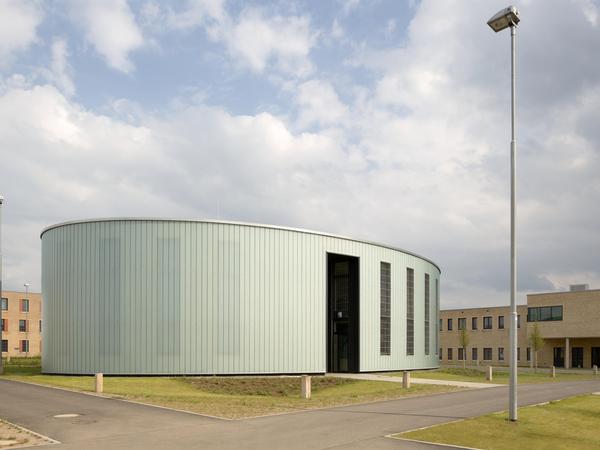 Rundes, eingeschossiges Kirchengebäude im Zentrum der Anlage mit hellgrüner Fassade, sehr geschlossene Fassade. Überhöhte Öffnungen, umgeben von Grünfläche. Im Vordergrund Wegekreuzung und ein Lichtmast. Im Hintergrund weitere Gebäude der Anlage und ein stark bewölkter, aber freundlicher, Himmel.