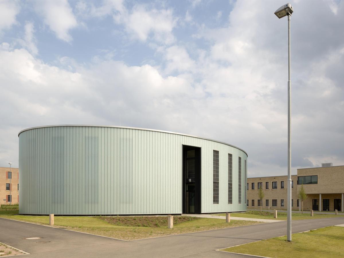 Rundes, eingeschossiges Kirchengebäude im Zentrum der Anlage mit hellgrüner Fassade, sehr geschlossene Fassade. Überhöhte Öffnungen, umgeben von Grünfläche. Im Vordergrund Wegekreuzung und ein Lichtmast. Im Hintergrund weitere Gebäude der Anlage und ein stark bewölkter, aber freundlicher, Himmel.