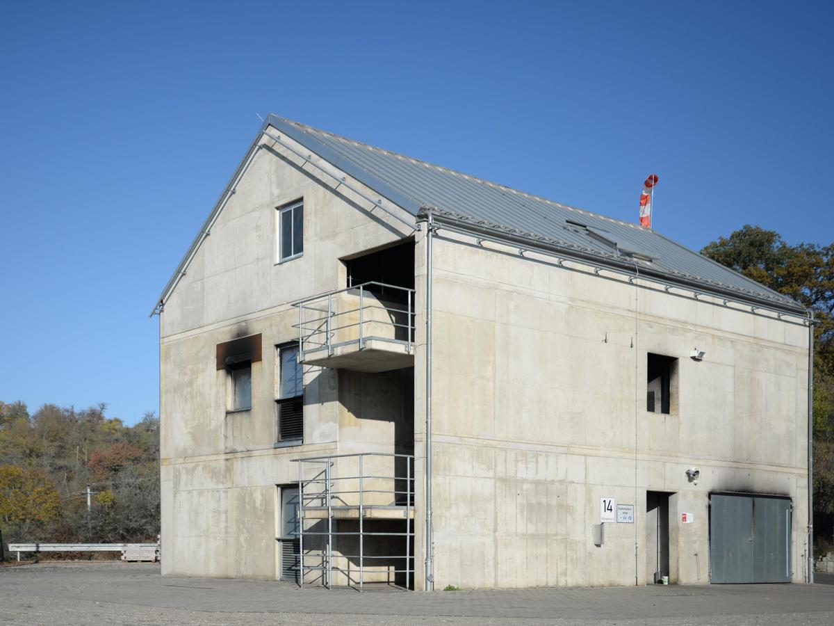 Gebäude aus Beton mit wenigen Fensteröffnungen mit und deutlichen Rußflecken, graues Satteldach. Im Hintergrund Hecken, Sträucher und kleine Bäume bei strahlend blauem Himmel.