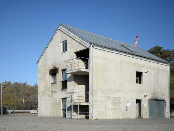 Gebäude aus Beton mit wenigen Fensteröffnungen mit und deutlichen Rußflecken, graues Satteldach. Im Hintergrund Hecken, Sträucher und kleine Bäume bei strahlend blauem Himmel.
