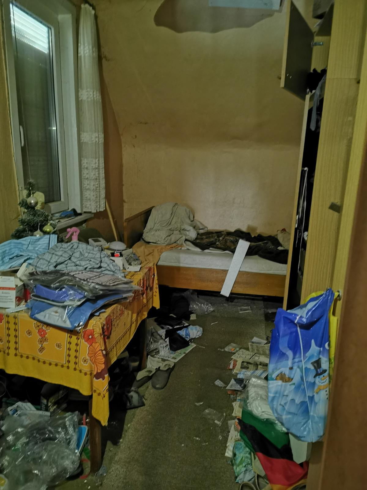vermülltes Zimmer; Bett unter Dachschräge, ein Fenster, alles verschmutzt und verschlissen
