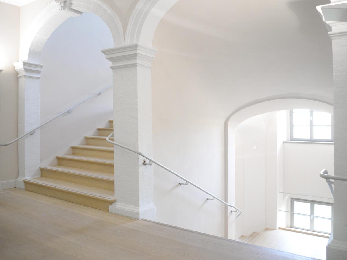 Treppenpodest mit Blick auf die dreiläufige Treppenanlage. Jeder Treppenlauf wird zu Beginn und Ende mit einem Rundbogen mit Schlussstein eingefasst. In der Mitte eine massive gemauerte Wand. Alle Wände hochweiß gemalert, ohne nenneswerte Zierde. Einfacher Stahlhandlauf mit Handlaufträgern. Am Zwischenpodest sind zwei Fenster mit grauen Rahmen erkennbar. Der Fußboden, wie auch die Treppenstufen, ist mit Holz belegt.