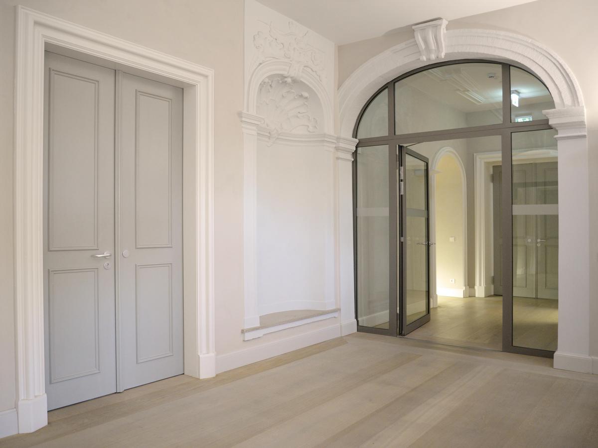 Großzügiges Treppenpodest mit grauer zweiteiliger Eingangstür mit Kassettierung und weißer profilierter Zarge. Lichtgraue Wände. Nach rechts führt eine Glastür in den Flur des Geschosses. Rundbogen in weiß.