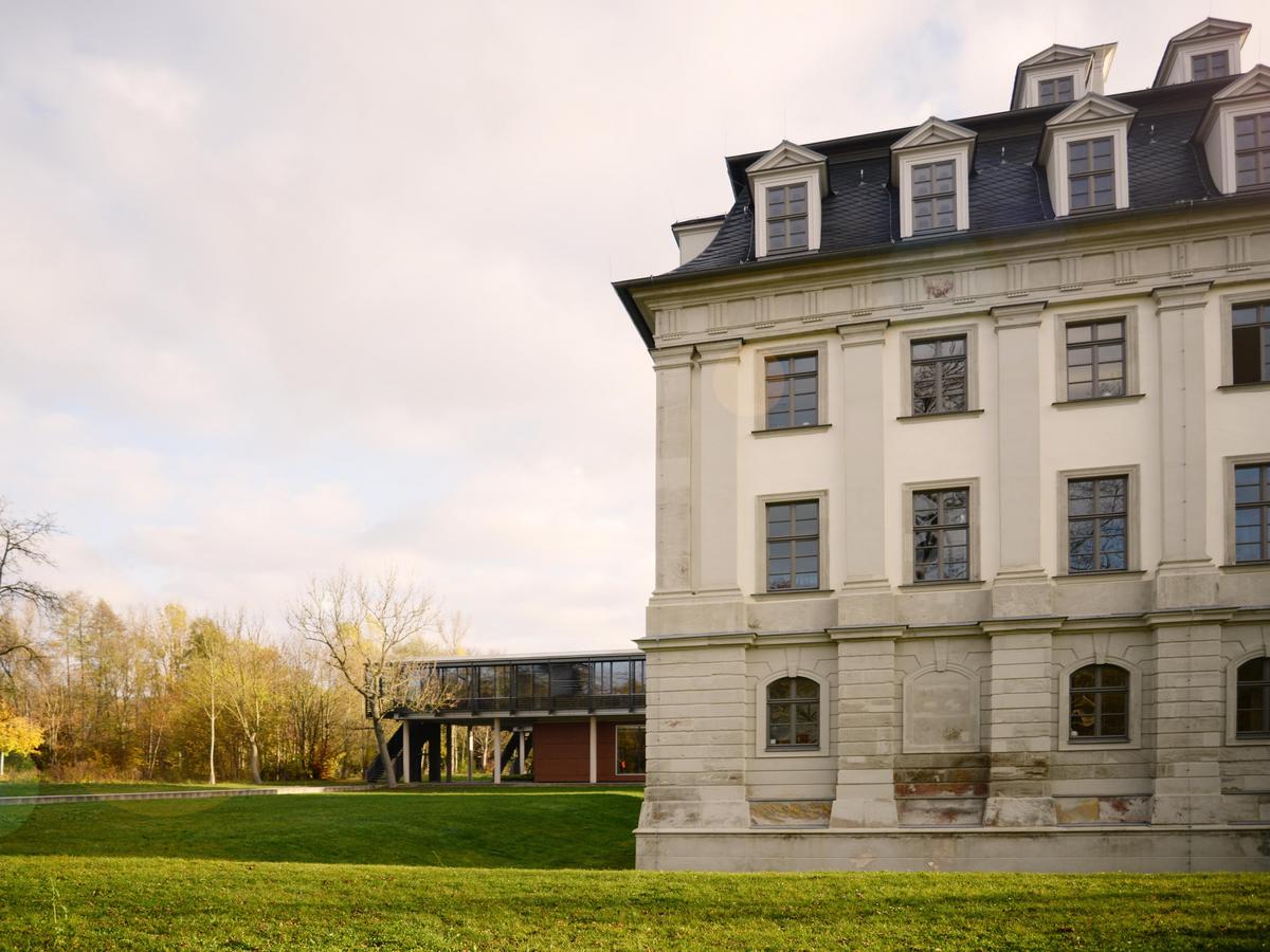 Halbe Westfassade des Barockschlosses mit durch Bäumen verdeckter Neubau im Hintergrund. Warmes Licht, leichte Bewölkung. Im Vordergrund eine grüne Wiese. Die Fassade ist nun detailreicher erkennbar. Rechteckige Fenster im 1. und 2. Obergeschoss, im Erdgeschoss Fenster mit Rundbogen. Sechs Gauben sichtbar, 4 unten, 2 oben.