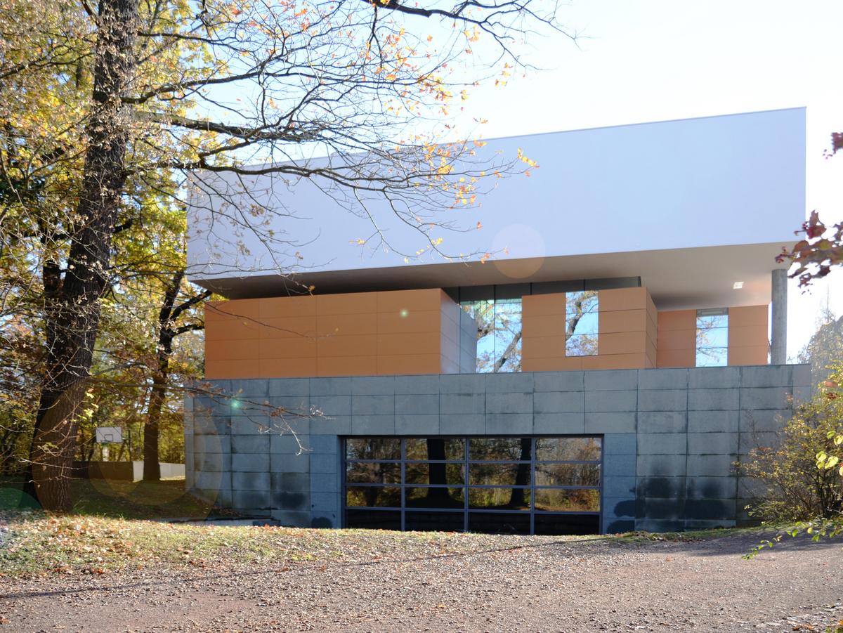 Kiesweg im Vordergrund, links ein riesiger Baum, rechts Gebüsch in Bild ragend. Im Zentrum das Schulgebäude als Neubau in seiner dreiteiligen Schichtung. Unten graue Natursteinplatten mit großen Fensterausschnitt, in der Mitte die orangenen Klassenräume, oben der weiße Kubus ohne Fenster.