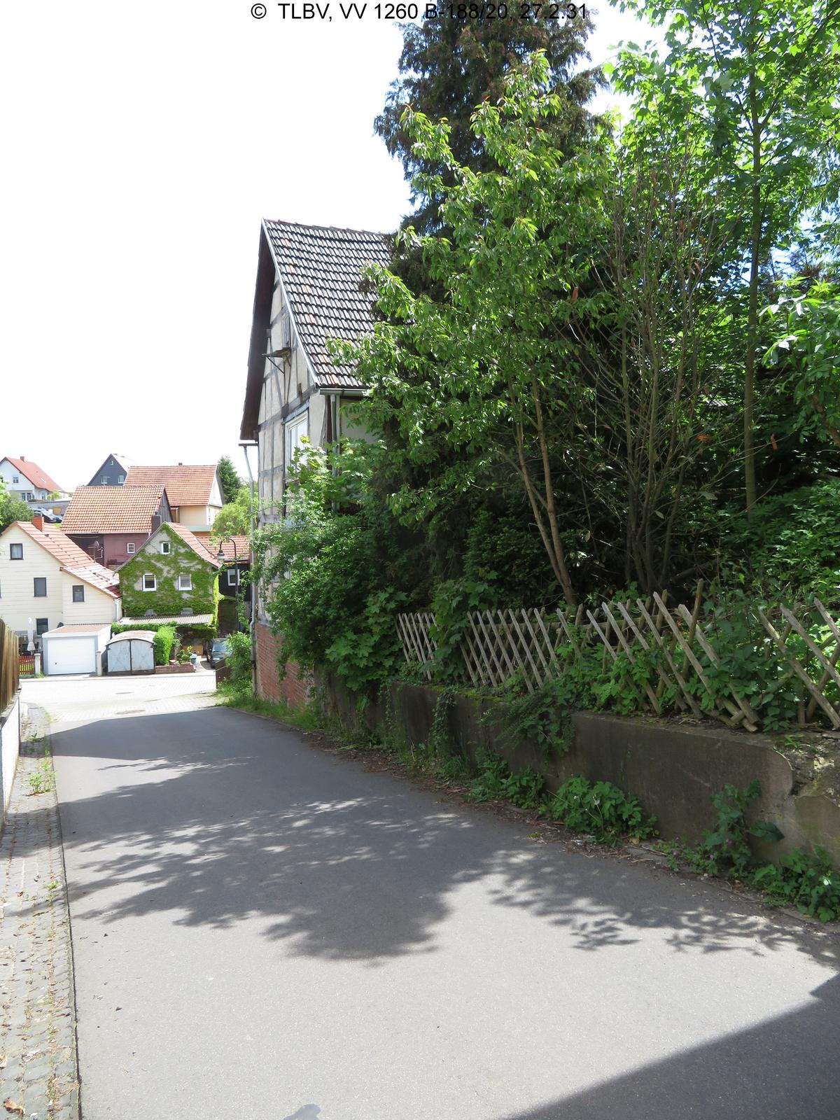 Bick die Straße entlang, rechts im Vordergrund zugewucherter Garten, im Anschluss giebelständiges Fachwerkhaus