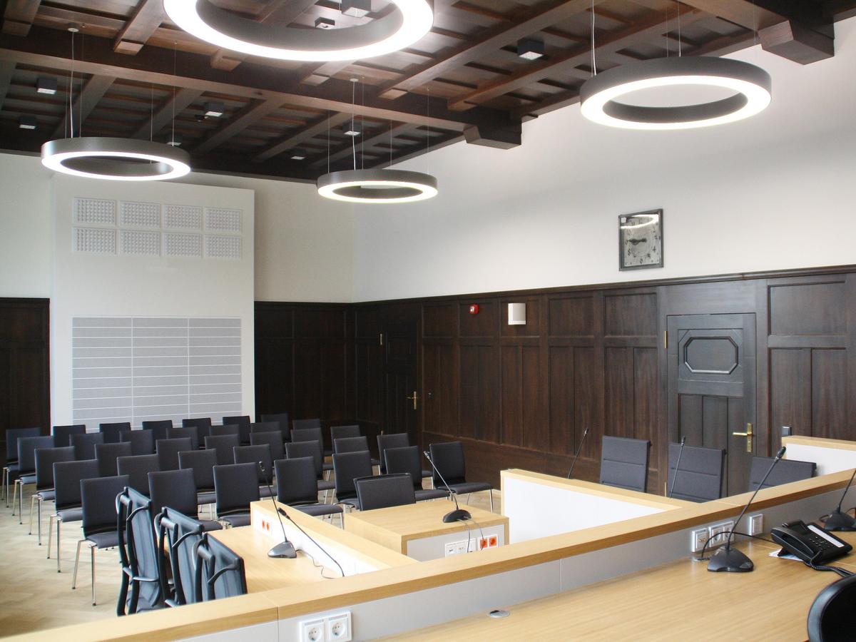 Innenfoto des Sitzungssaals aus Blickrichtung des Richters. Schwere dunkelbraune Holzkonstruktion an der Decke, hohe Raumhöhe, abgehangene kreisrunde helle Deckenleuchten. Sitzplätze für Zuschauer auf schwarzen Stoffsitzen. Sitzplätze für die Gegnerparteien im Bildzentrum. Stirnwand mit weißem Technik-Panel.
