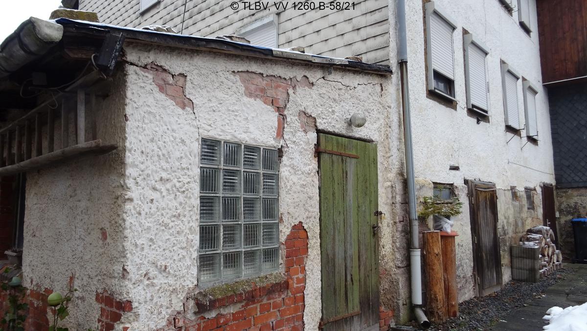 Detalansicht auf eingeschossigen Anbau mit Pultdach, Fenster aus Glasbausteinen und Schuppentür aus Holz; erhebliche Putzabblätterungen