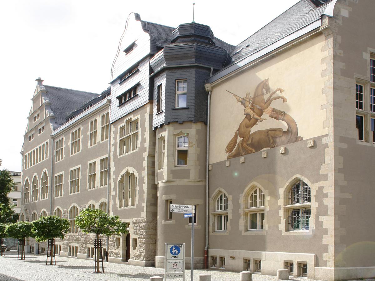Fassade mit eindrucksvollem großen Wandgemälde vom Heiligen Georg im Kampf mit einem feuerspuckenden Drachen. Im Vordergrund ein menschenleerer Platz mit grauem feinen Pflaster. Durch Schatten zeichnet sich die Gebäudekubatur anschaulich ab.