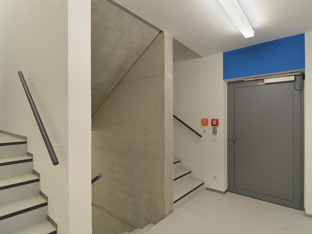 Funktionstreppenhaus, d.h. sachliche Gestaltung, Betonwände, außer im Treppenaugen weiß geputzt und gemalert. Einfacher Rundrohr-Handlauf in Dunkelgrau. Drei Treppenläufe reihen sich nebeneinander. Rechts ist eine schwere graue, aber moderne, Tür zu Erkennen. Darüber eine große blaue Fläche, die auf das blaue Magazin hindeutet.