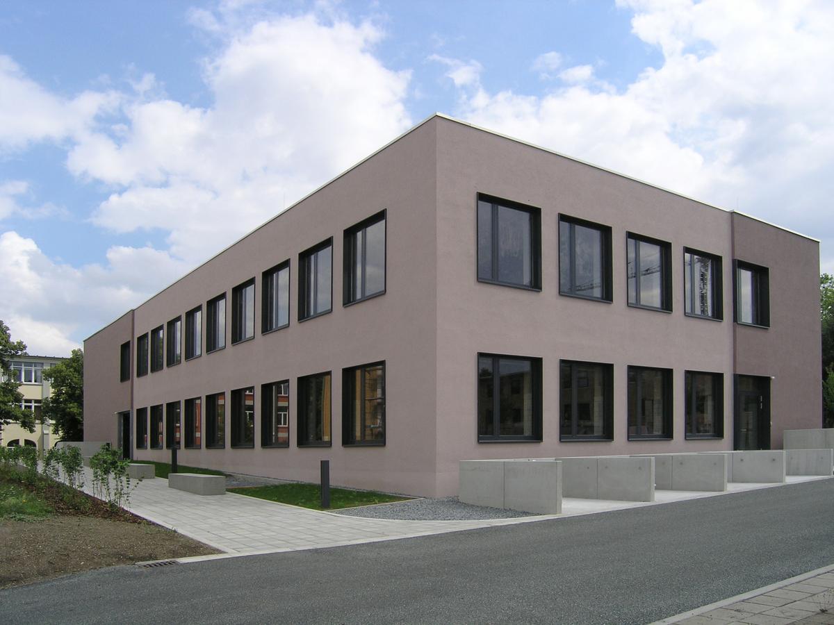 Ein braunes Gebäude mit schwarzen Fenstern, davor führt eine Straße vorbei. Die Fenster sind stark gerastert. Links führt ein Gehweg zur Vorderseite des Gebäude. Ein klarer Tag mit leichter Bewölkung.