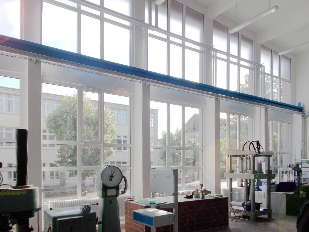 Zweigeschossiger Versuchsraum mit Glasfassade, viel einstrahlendes Sonnenlicht. Schwarze Lüftungsrohre an der Rückwand. Die blaue Krananlage thront über dem Raum und wirkt schwer und dennoch schwebend.