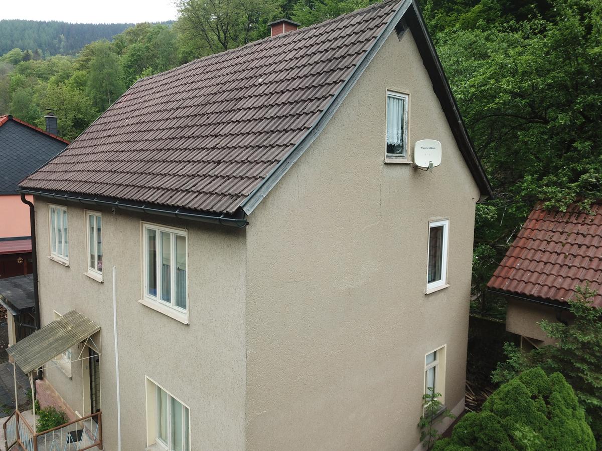 Blick auf die Giebelseite des zweigeschossigen Wohnhauses; im Giebel sowie je Etage befinden sich je ein Fenster an der Giebelfront; hinter dem Haus schließt sich direkt der Hang mit Baumbewuchs an