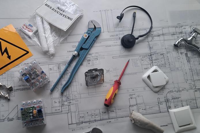 Auf einem ausgebreiteten technischen Gebäudeplan aus Papier liegen verschiedene Werkzeuge und Zubehörteile, die die Vielfalt das Themas technische Gebäudeausstattung symbolisieren.