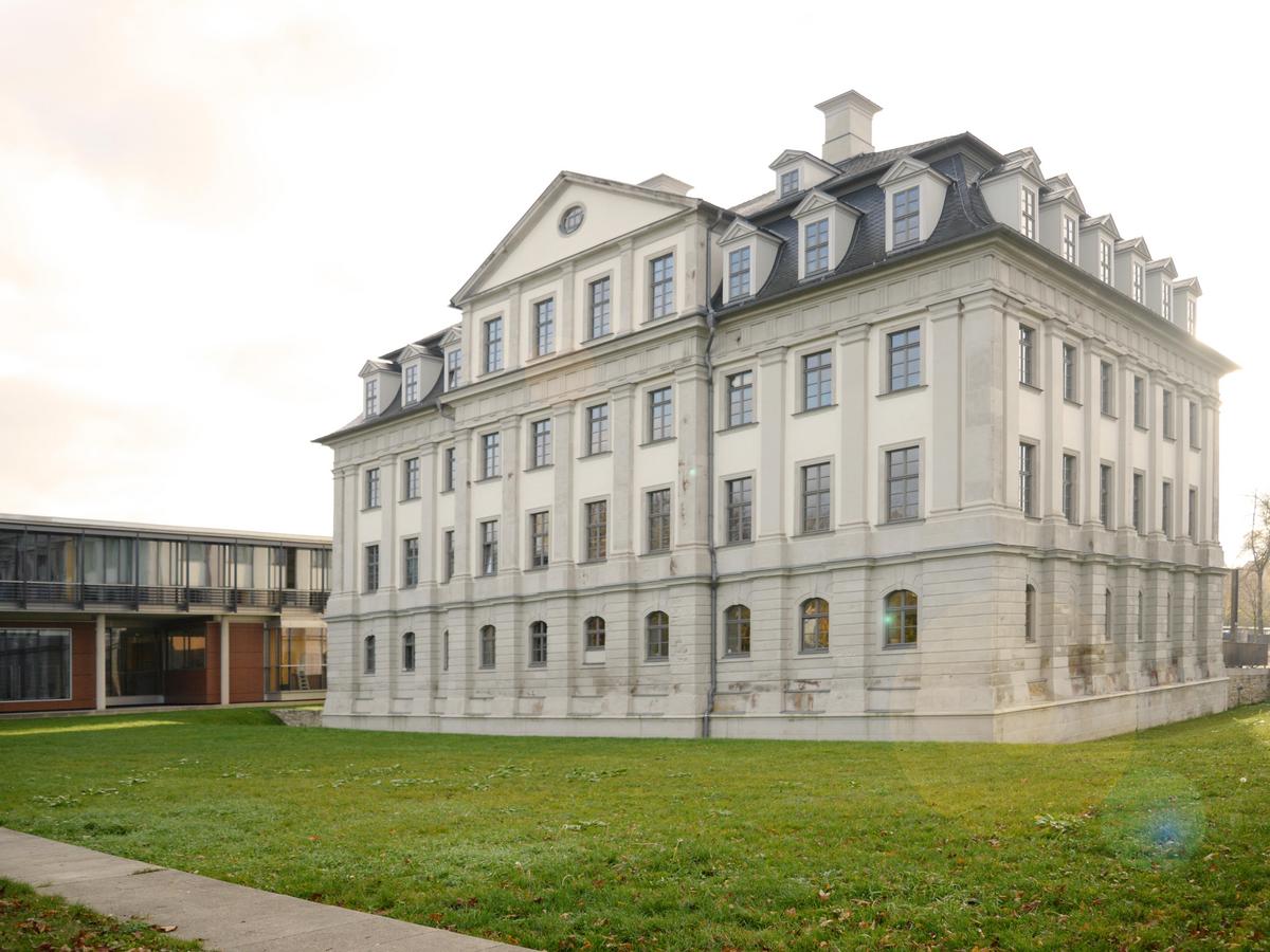 Schloss Tinz im Zentrum des Bildes. Dreigeschossiges Barockschloss mit aufragenden Giebel, grauer Schieferdeckung, vielen Gauben und Schornsteinen. Leichte Bewölkung, die Sonne versteckt sich hinter dem Schloss. Im Vordergrund läuft ein schmaler Weg nach links aus dem Bild. Grüne Wiese ohne Blumen. Links im Hintergrund der zweigeschossige Neubau der Dualen Hochschule, flach und abgeduckt.