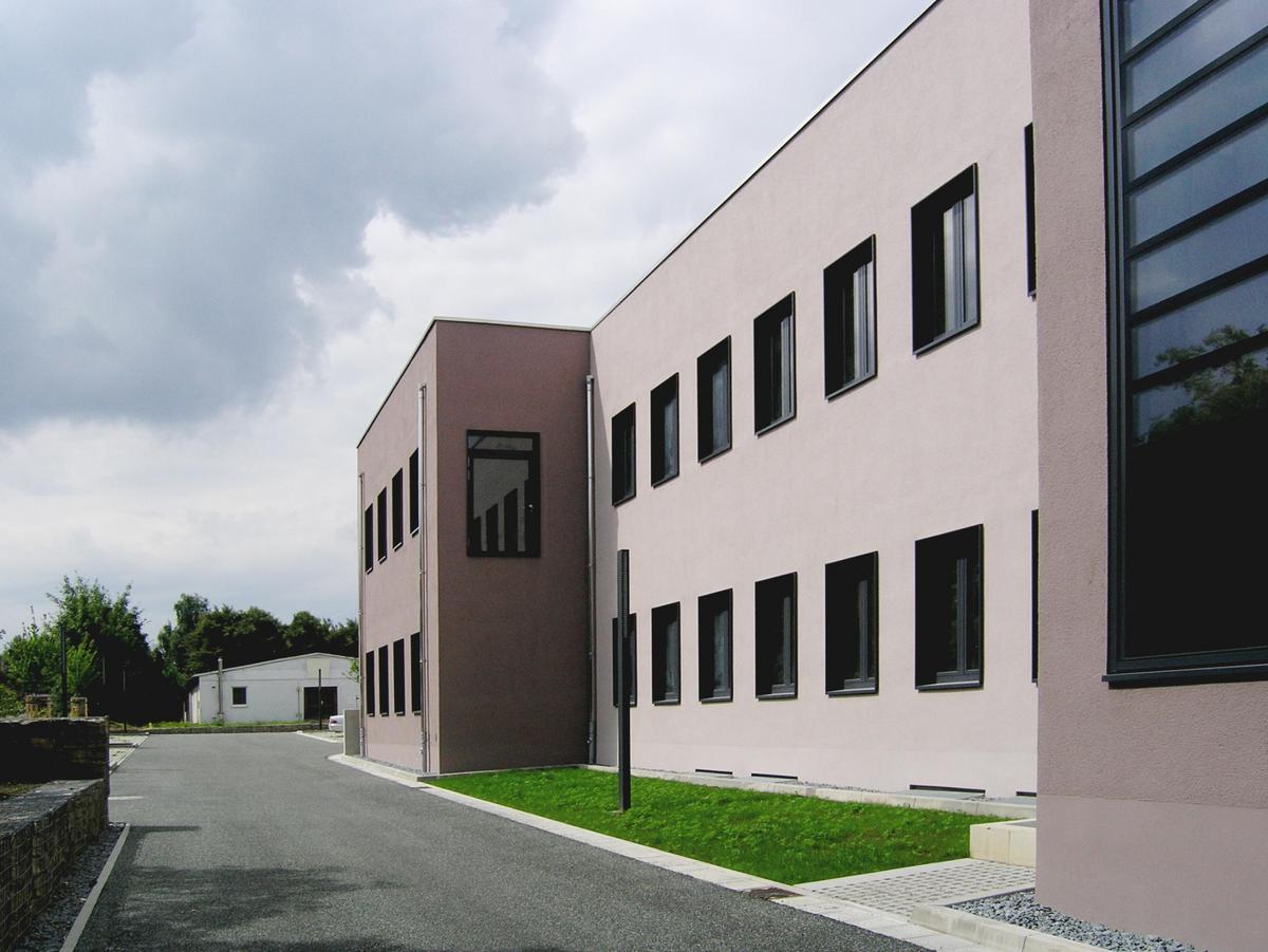 Schmale asphaltierte Straße führt am Betonbau vorbei zur Rückseite, man erkennt eine Dreiteilung der Fassade. Eine kleine Grünfläche liegt vor dem zurückspringenden Gebäudeteil. Starke Bewölkung, im Hintergrund sieht man Bäume und eine flache Lagerhalle.