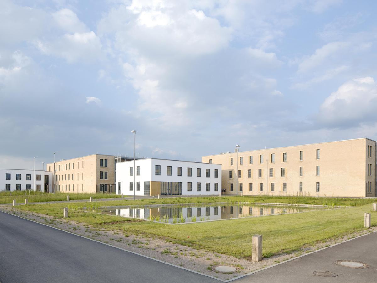 Perspektive mit Löschteich und kreuzenden Asphalt-Wege im Vordergrund, bewölkter aber freundlicher Himmel. Zwei zweigeschossige weiße Gebäude im Bildzentrum, denen dahinter jeweils ein sandsteinfarbenes dreigeschossiges Gebäude angegliedert ist.