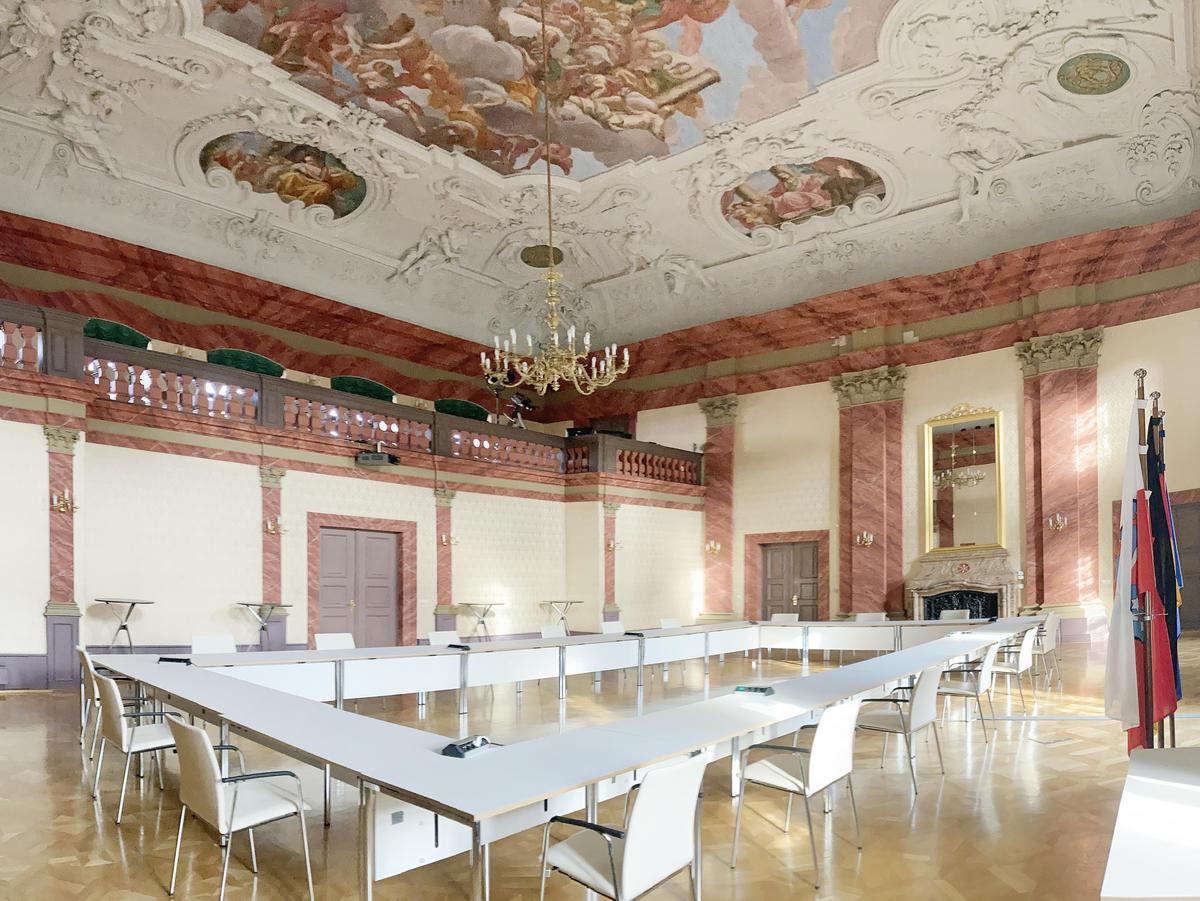 Barocksaal mit imposanten Deckenfresko. Stuck an jeden freien Quadratzentimeter Fläche in weiß. Die Wände werden von roten Marmorsäulen gehalten. Parkettfußboden. In der Raummitte Tische und Stühle in weiß in Form einer Null aufgestellt. Rechts stehen Fahnen die schlaff nach unten hängen. Links ist eine Galerie in der zweiten Ebene zu erkennen. Davor stehen vier Stehtische und dazwischen die zweiflügelige Eingangstür in grau.