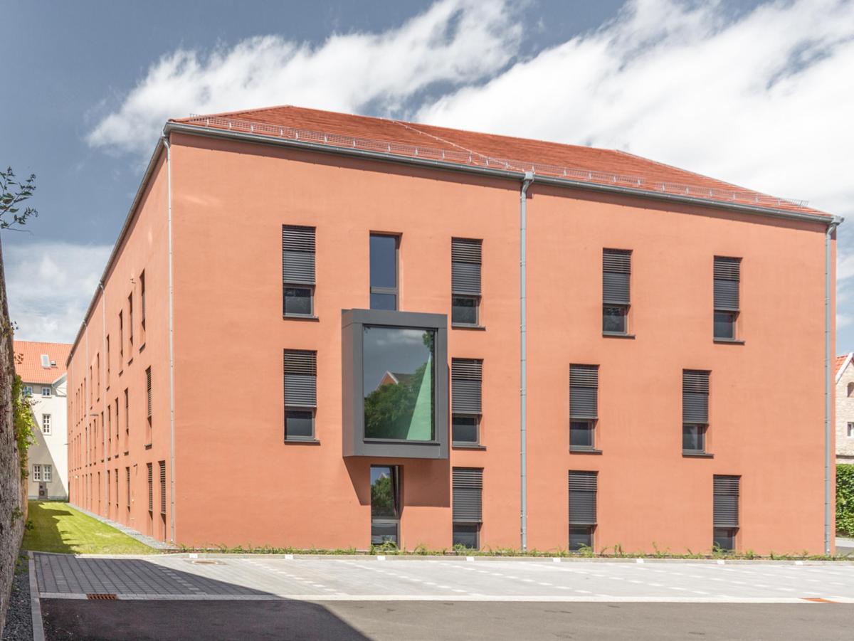 Dreigeschossiges orangenes Gebäude mit schwarzen Fenster und roten Satteldach. Links ist eine alte Stadtmauer die in das Bild hineinläuft. Im Vordergrund eine Straße mit rechts und links Stellplätzen. Ein heiterer blauer Himmel mit etwas Wolken.