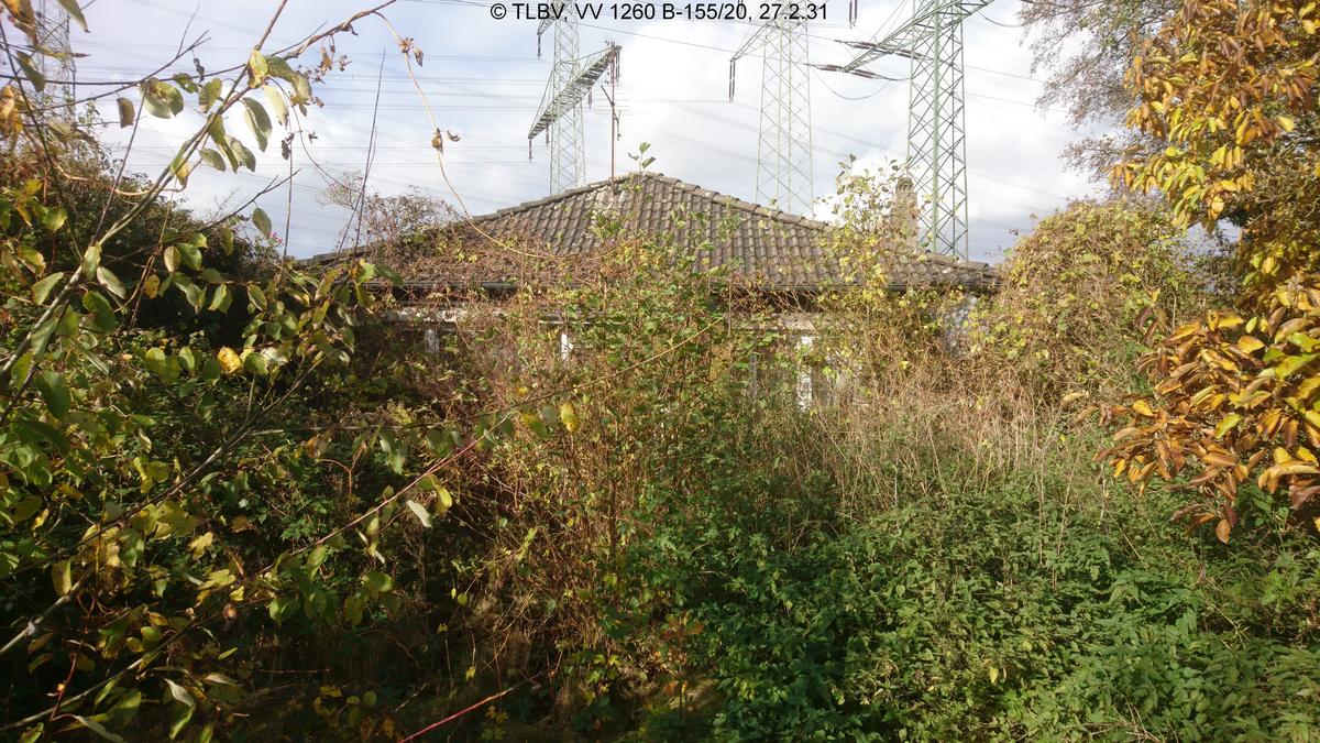 über stark verwilderten Blick auf den Wohnbungalow, hinter dem Haus drei Starkstrommasten für Hochspannungsleitungen
