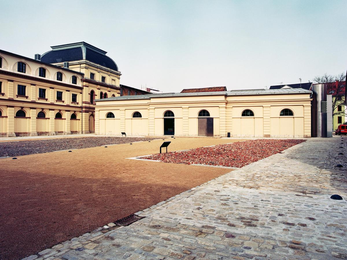 Ein großer Innenhof mit vier verschiedenen Bodenbelagsarten. Bruchstein aus Travertin, wassergebundene Decke eingerahmt vom Travertin und frei platziert zwei rechteckige Flächen mit Bauschutt. Das ist das Kunstwerk Zermahlene Geschichte. Im Hintergrund der Westflügel des Marstalls, rechts davon das flache Gebäude, die ehemalige Reithalle. Alle Gebäude sind sandsteinfarben mit roten Akzenten.