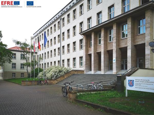 Straßenperspektiven, vorne links ein gepflasteter Weg, das Gebäude hat eine breite Treppenanlage bevor die Eingangssäulen erreicht werden. Daneben Grasflächen und ein weißes Schild mit TMUEN-Beschriftung, im Vordergrund stehen vier Fahrräder an den Ständern, das Gebäude ist eierschalenweiß, viergeschossig. Man sieht ein Staffelgeschoss im Ansatz.