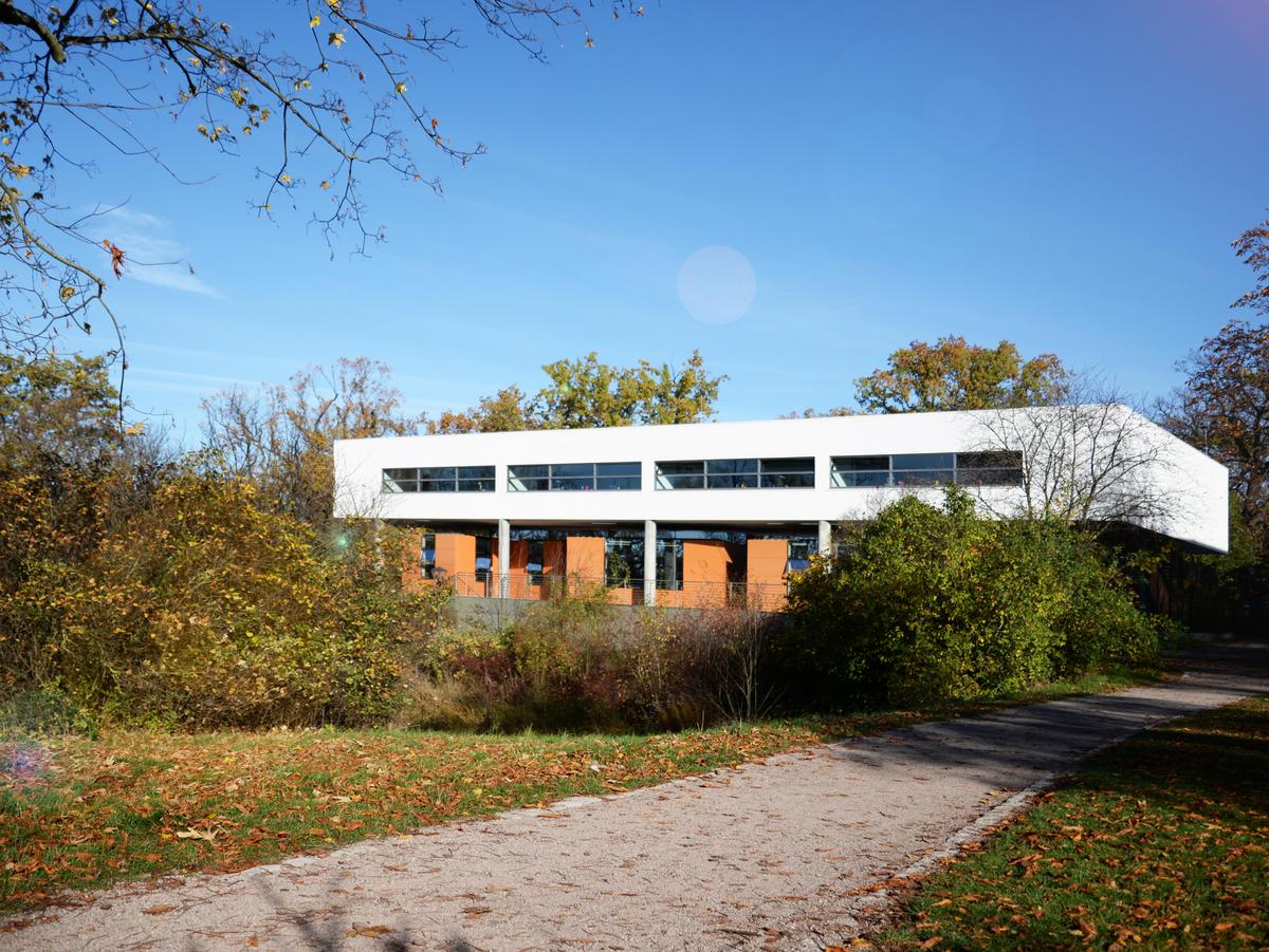Ein Kiesweg führt zum Neubau, rechts und links Grünpflanzen und Äste ragen in das Bild. Der Neubau wird teilweise von Gebüsch verdeckt. Der flache weiße Neubau strahlt im Sonnenlicht. Die orangenen Boxen der Glasfassade sind gut zu erkennen. Blauer Himmel, Sonnenschein.