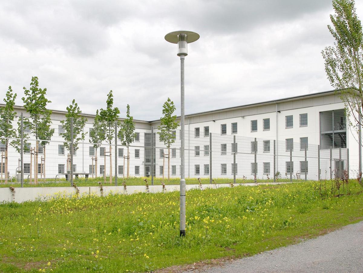 Ein Foto mit angeschnittenen Weg unten rechts, anschließend eine Wiese mit gelben Blumen, dann folgt ein Zaun auf massiven Betonfertigteilen. Hinter dem Zaun ist das L-förmige Zellengebäude zu sehen. Eine einfach weiße Lochfassade. Das L umfasst einen Innenhof mit jungen Bäumen in ihren Wachsgestellen.