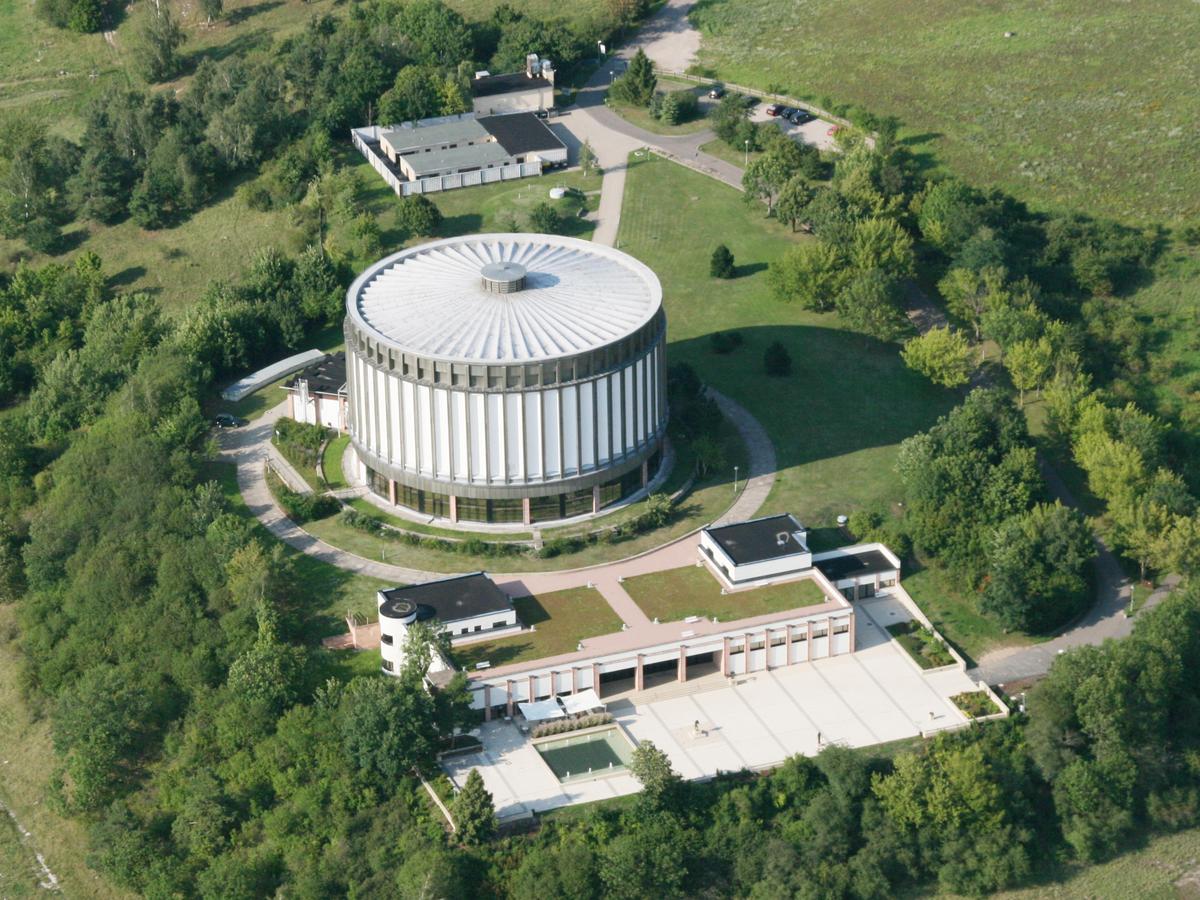 Luftaufnahme vom Schlachtberg, im Vordergrund der Vorplatz, von rechts führt der Weg auf den Platz. Vom Vorplatz geht es in den eingeschossigen Vorbau. Man schaut auf das Dach des Rundbaus, es wirkt wie Speichen mit Randaufkantung. Umgeben ist das Gelände von grünen Wiesen und kleineren Ansammlungen von Bäumen und Sträuchern.