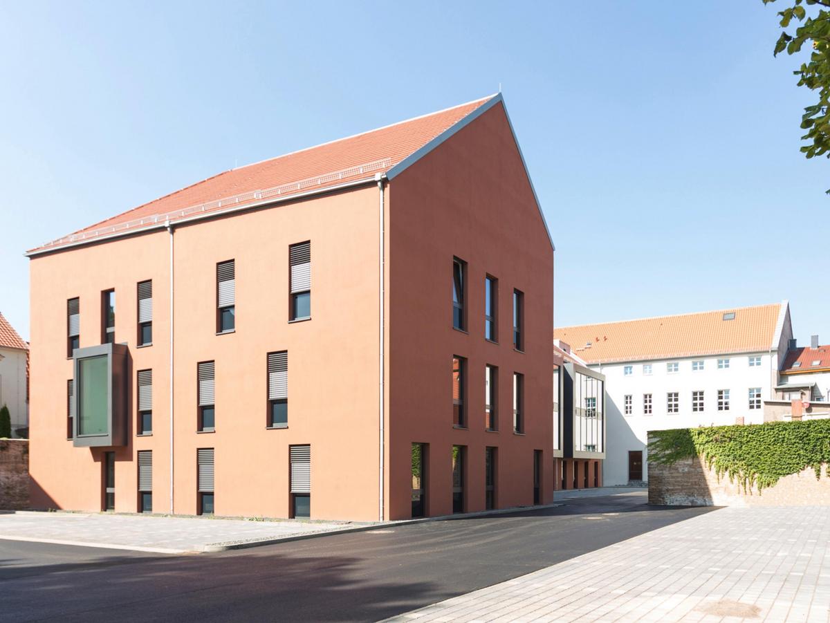 Im Vordergrund jede Menge Parkplätze, rechts die alte Stadtmauer die in das Bild hineinläuft. Im Hintergrund das alte weiße Amtsgericht. Im linken Vordergrund die Südansicht des dreigeschossigen orangenen Anbaus mit den schwarzen Fenster, die Giebelseite liegt im Schatten.