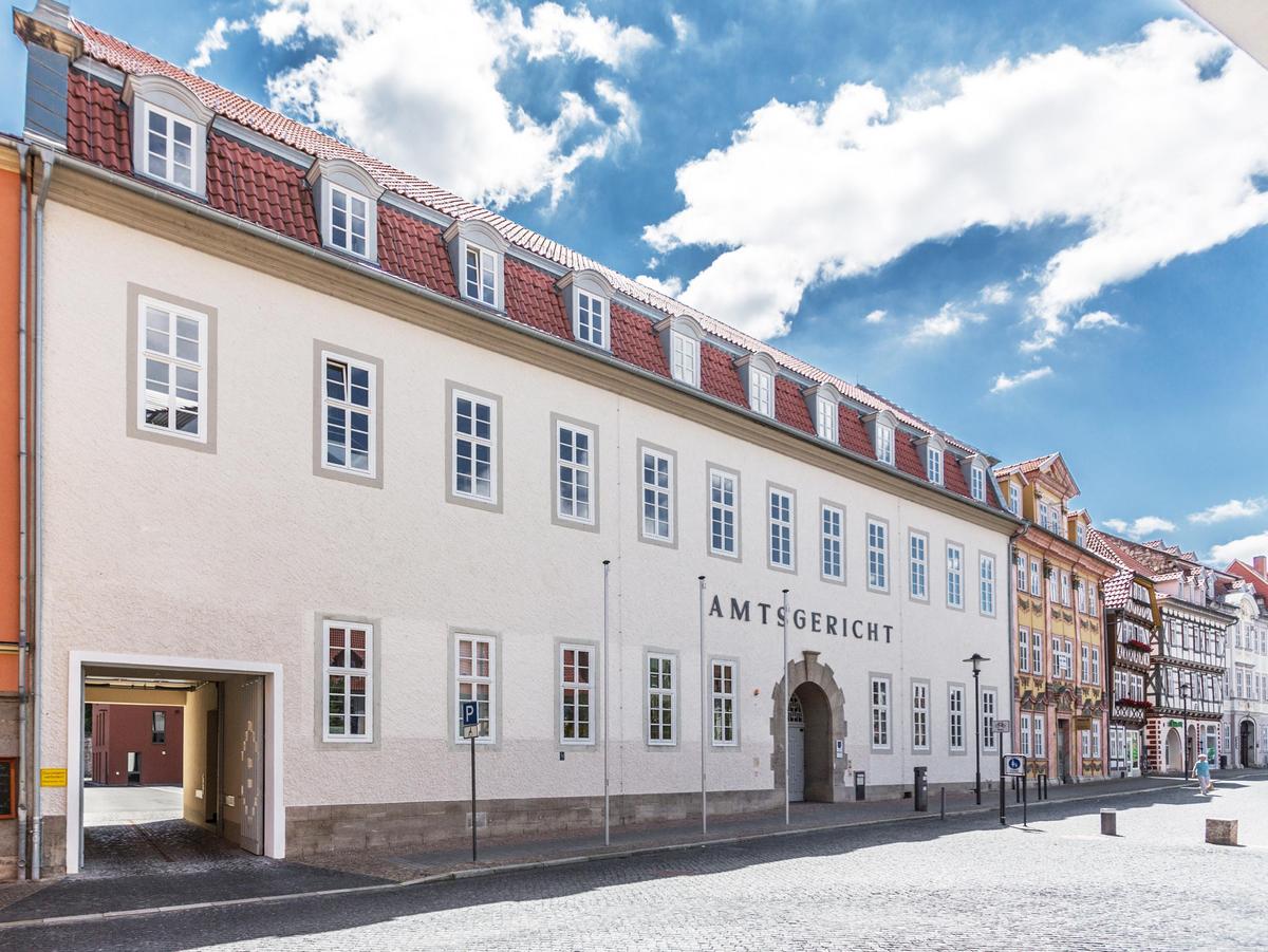 Blick auf den Altbau am Untermarkt. Zweigeschossiger Bau mit Mansarddach mit roten Dachziegeln und 10 Dachgauben in weiß mit Tonnendach. Alle Fenster streng gerastert angeordnet mit Sprossenteilungen. Der Haupteingang ist durch den Rundbogen gut zu erkennen. Ein großer Schriftzug "Amtsgericht" darüber. Die Einfahrt links lässt den rückwärtigen neuen Anbau erahnen. Vor dem Gebäude drei leere Fahnenmasten und ein menschenleerer Untermarkt. Eindrucksvolle Wolken am blauen Himmel.