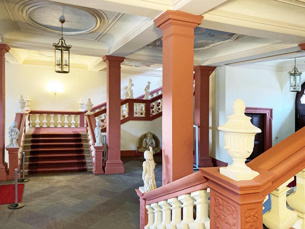 Foyer, die Farben der Fassade spiegeln sich hier wieder. Mächtige, schwere, massive Treppengeländer mit weißen Rundsäulen als Verstäbung. Schwarze Fliesen auf dem Boden. Marmor-Skulpturen von Menschen oder Gefäßen an den Knickpunkten der massiven Geländer. Kassettierung der Decke mit Stuck in den Feldern in weiß.
