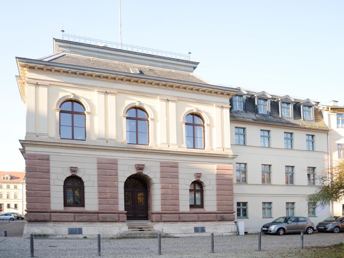 Westfassade des Westresalits mit deutlich massiverer Formsprache. EG mit Eingangstür mit dunklen Holz, rechts und links jeweils große Fenster mit Rundbögen und Sprossen. Im OG nur noch hellgelb und reduziertere Fassade und drei gleichmäßig verteilte Rundbogenfenster. Darüber das Dach mit Dachterrasse. Rechts direkt anschließend der spätere Anbau in sachlicher Formsprache und unterschiedlicher Geschosshöhe. 4 Geschosse im Vergleich zu 2 vom Westflügel. Davor eine gepflasterte Straße mit parkenden Autos. Rechts geht die Häuserfront weiter. Ein angeschnittener Baum ragt ins Bild.