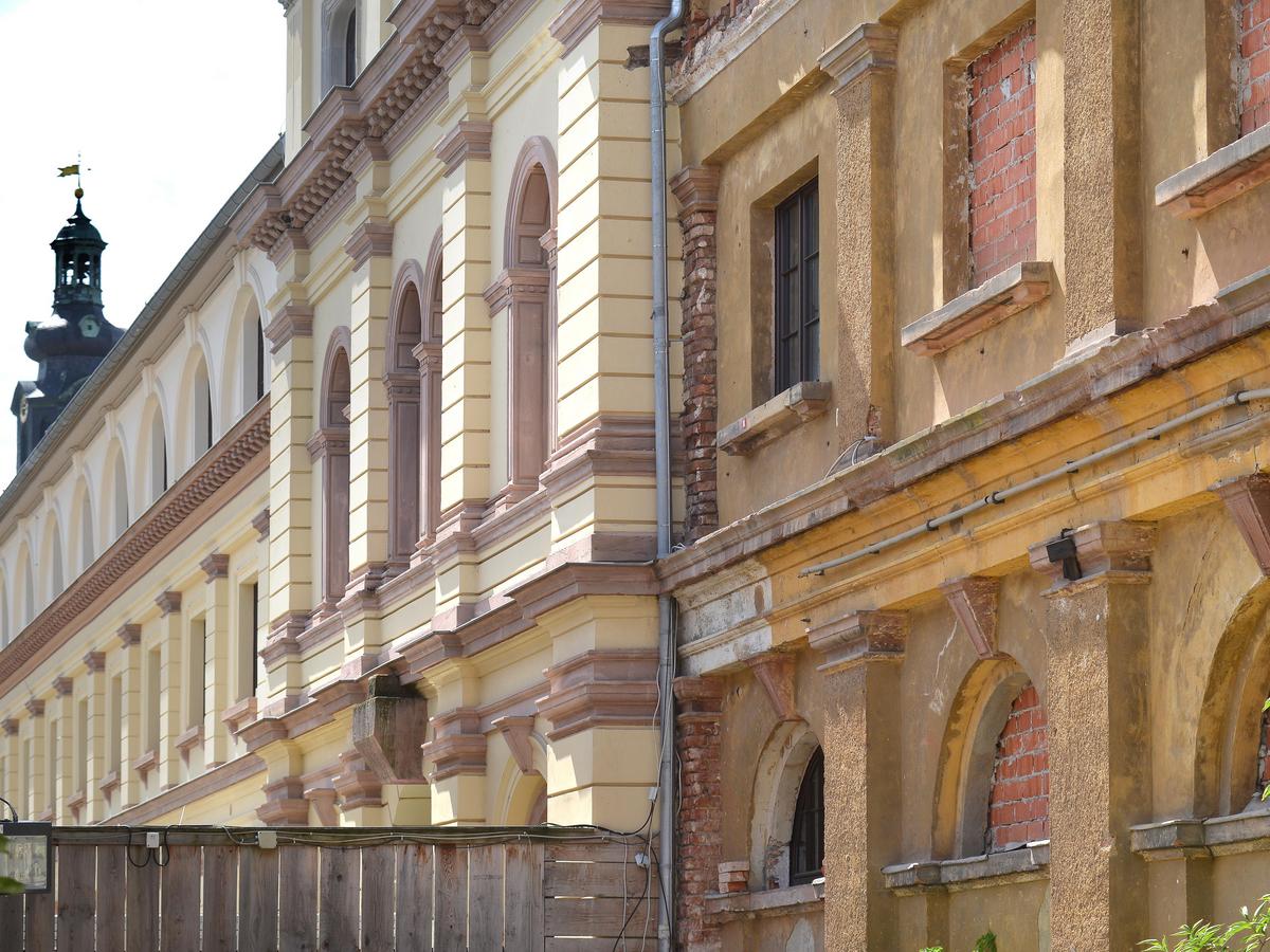 Detailfoto einer Fassade des Westflügels. Harter Kontrast: rechte Bildhälfte unsaniertes Gebäude mit wild gemauerten Fensteröffnungen, herunterhängenden Kabeln, wild verlegten Kabeln und bröckelnden Putz. Dazwischen ein Holzzaun als Sichtschutz, dahinter der sanierte Bereich des Marstalls ab Mittelrisalit. Sandsteinfarben, sauber, mit dunkelroten Akzenten.