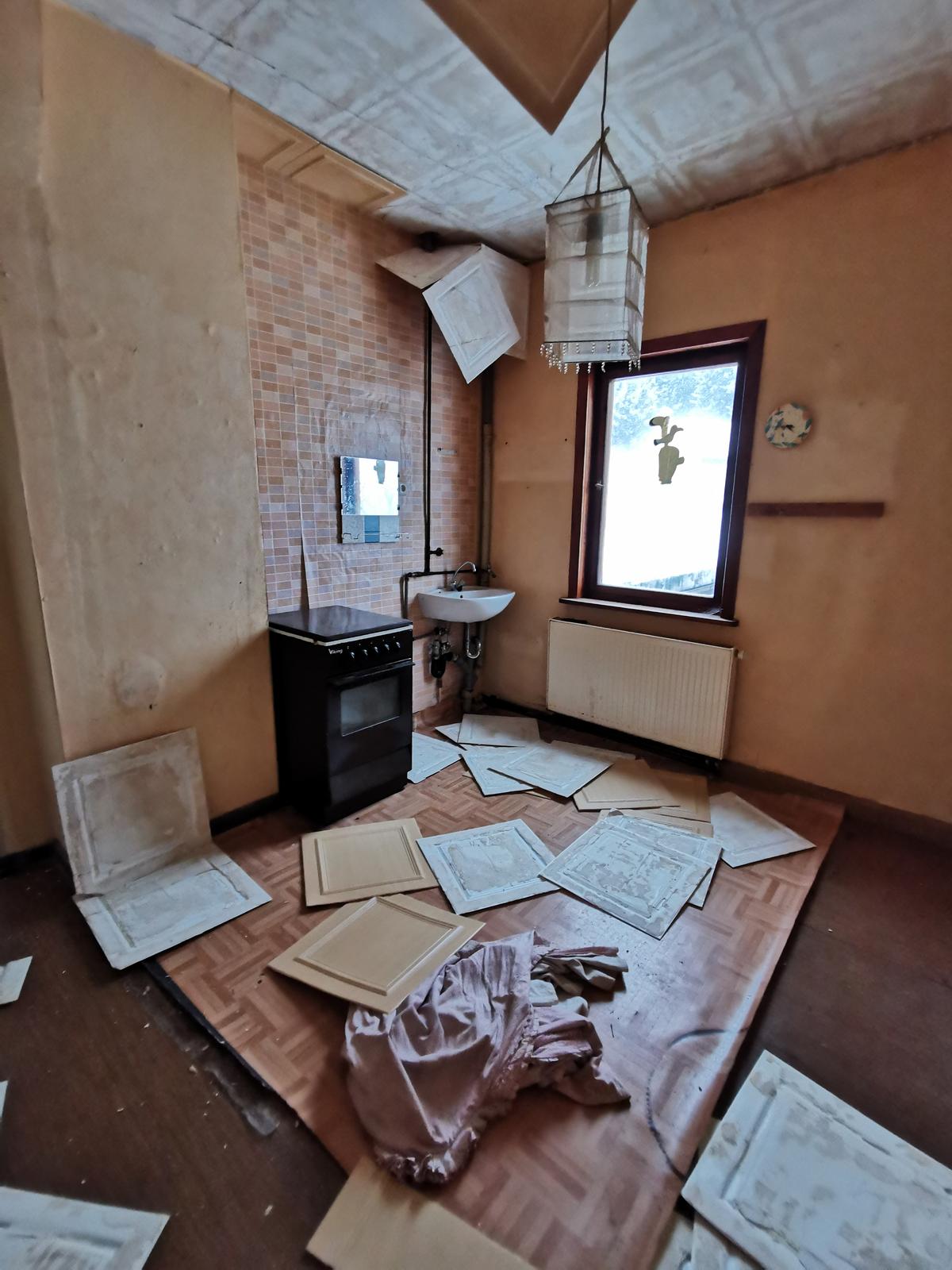 Zimmer mit gewelltem Fußbodenbelag und herabfallenden Deckenplatte, 1 Waschbecken, ein Herd, ein Fenster, ein Heizkörper
