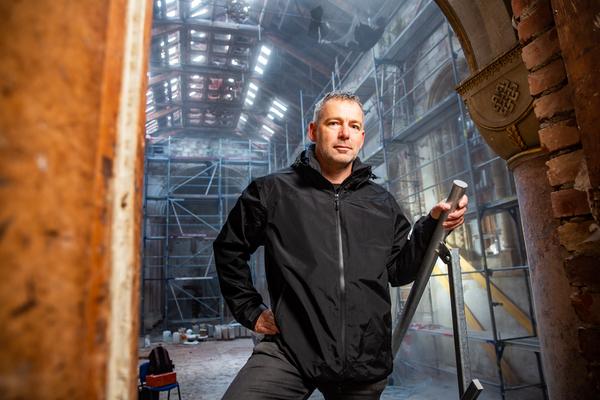 Junger schwarz-gekleideter Mann in historischem Türbogen; im Hintergrund ein eingerüsteter Kirchenraum