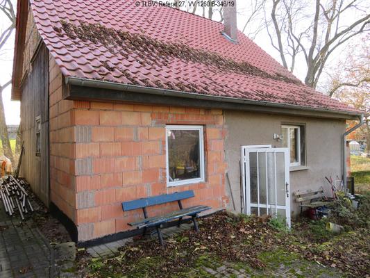 straßenseitige Ansicht eine kleinen Einfamilienhauses, 1 Geschoss plus Satteldach, mittig die Hauseingangstür, recht und links davon je ein Fenster, eine Haushälfte ist unverputzt