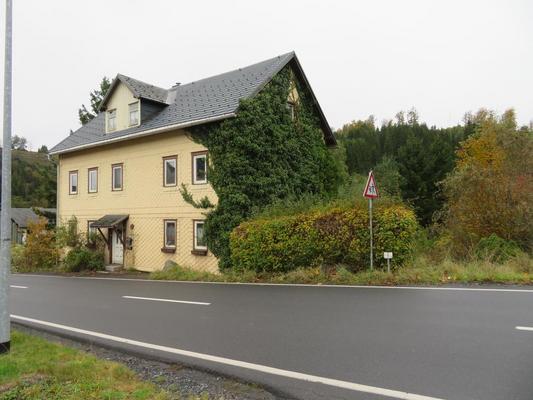 ein zweigeschossiges Huas, parallel zur Landstraße mit gelben Fassadenschindeln; Giebelseite von wildem Wein bewachsen