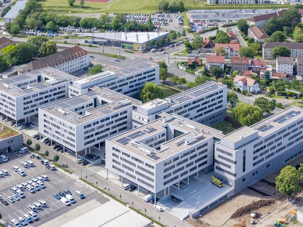 Luftaufnahme: vorne links Parkplätze mit diversen Streifenwagen und Transportern. Der enorme bammartige Neubau ist zu sehen. Drei Hofhäuser, rechteckig mit jeweils einen rechteckigen Innenhof. Alle Fassaden weiß. Man sieht dunkle Fensterbänder. Im Hintergrund die Kranichfelder Straße, 4-spurig, Wohnbebauung und eine Tankstelle unter einem dreieckigen Dach.