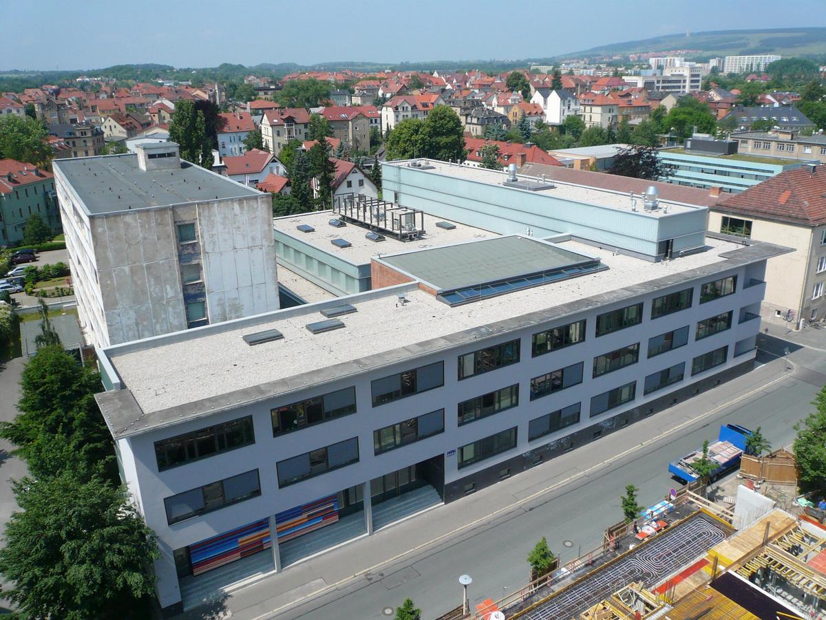 Fassadenansicht von einem gegenüberliegenden Gebäude mit der Straße im Vordergrund von links unten nach rechts mittlere Bildhöhe verlaufend. Das Gebäude ist E-förmig, alle Gebäudeteile mit unterschiedlichen Höhen und Flachdächern. Keine Personen auf der Straße. Im rechten unteren Bilddreieck ist eine Baustelle zu erkennen. Im Hintergrund die Stadt Weimar mit den historischeren Gebäude und rot gedeckten Dächern. In der Ferne das Mahnmal vom KZ Buchenwald.